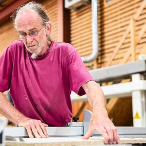 May include: A man wearing a pink t-shirt uses a table saw to cut a piece of wood. He is wearing glasses and has a beard.