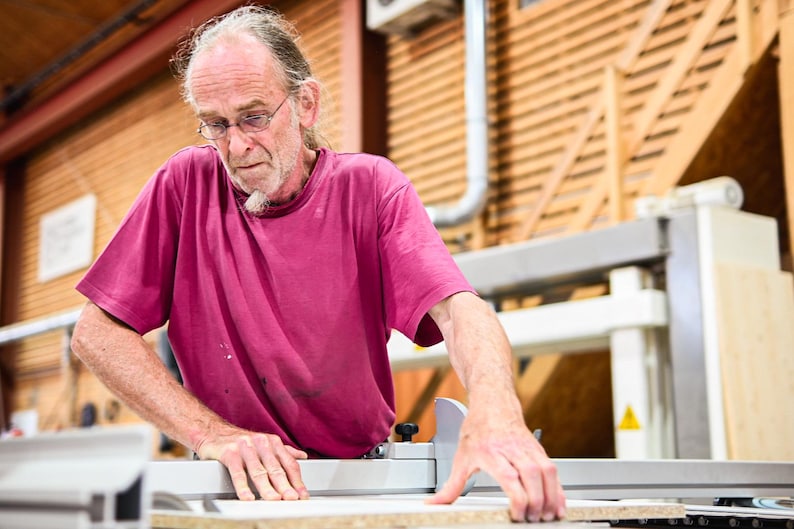 May include: A man wearing a pink t-shirt uses a table saw to cut a piece of wood. He is wearing glasses and has a beard.