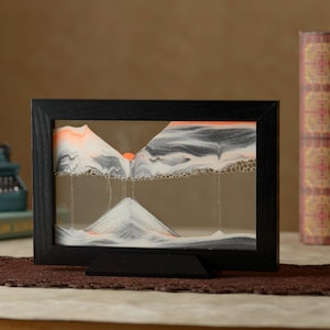 Peut inclure: Une image de sable encadrée en noir avec un motif de sable blanc et gris qui ressemble à des montagnes. Le sable coule vers le bas pour créer une chaîne de montagnes. L'art du sable est sur une surface brune.