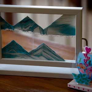 May include: A white frame sand art picture with a blue and white sand design. The sand art is of mountains and is in a white frame with a white base. The frame is on a wooden table.
