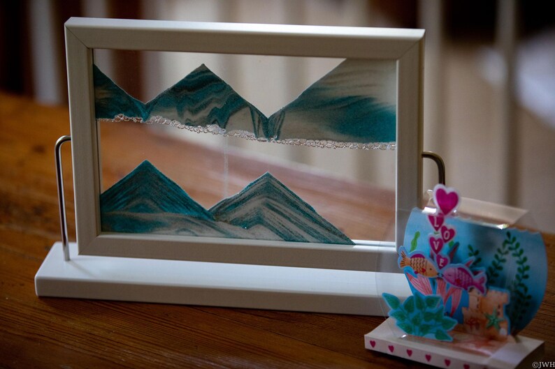 May include: A white frame sand art picture with a blue and white sand design. The sand art is of mountains and is in a white frame with a white base. The frame is on a wooden table.