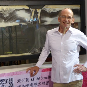 May include: A man in a white shirt stands in front of a framed sand art piece. The sand art is in shades of gray and white and features the text ".T.Я.". The man is smiling and has his hand on his hip. The text on the sign below the sand art reads "欢迎到 K11 Art Store (B) 逛购更多艺术精品".