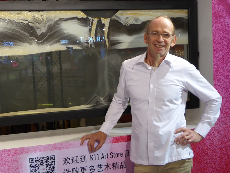 May include: A man in a white shirt stands in front of a framed sand art piece. The sand art is in shades of gray and white and features the text ".T.Я.". The man is smiling and has his hand on his hip. The text on the sign below the sand art reads "欢迎到 K11 Art Store (B) 逛购更多艺术精品".