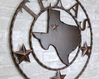 Könnte beinhalten: Eine Wanddekoration aus Metall mit der Umrandung des Bundesstaates Texas, wobei das Wort "TEXAS" in einem Kreis darum herum geschrieben ist. Die Umrandung des Bundesstaates ist mit Stacheldraht verziert und es gibt fünf Sterne, einen innerhalb der Umrandung des Bundesstaates und vier um den äußeren Kreis herum.