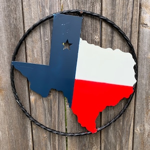 May include: A metal wall hanging in the shape of the state of Texas, painted in the colors of the Texas flag: navy blue, white, and red. A star is cut out of the blue section. The Texas shape is surrounded by a black, twisted metal ring.