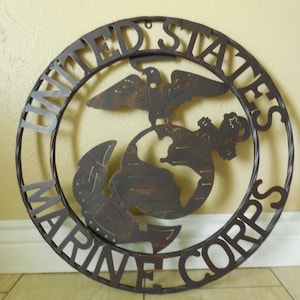 May include: A metal wall decor featuring the United States Marine Corps emblem. The emblem is a circle with the words "United States Marine Corps" around the outside. Inside the circle is a globe with an eagle perched on top of it. The eagle is holding a ribbon in its beak. The globe is surrounded by an anchor.