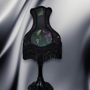 May include: A black table lamp with a floral patterned lampshade. The shade features a dark background with green and purple floral accents. The lamp has a black base and a fringe around the bottom of the shade.