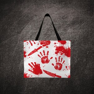 May include: White tote bag with a repeating pattern of red handprints, blood splatters, and knives. The bag has a black handle and is made of a durable material. The design is bold and eye-catching.