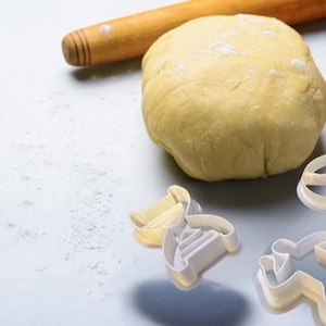 May include: A ball of dough sits on a white surface next to a wooden rolling pin and cookie cutters. The dough is lightly dusted with flour.