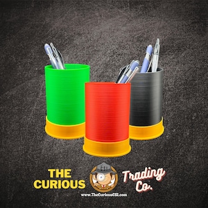 May include: Three cylindrical pen holders in green, red, and black, each with a yellow base. Silver pens are visible inside the holders. The Curious Trading Co. logo is at the bottom.