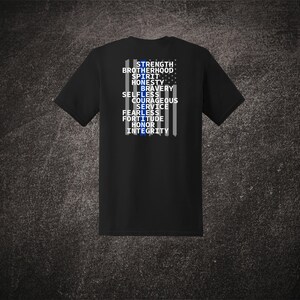 Może przedstawiać: Czarna koszulka z graficznym wzorem przedstawiającym niebiesko-białe ułożenie przypominające flagę. Projekt zawiera słowa "STRENGTH, BROTHERHOOD, SPIRIT, HONESTY, BRAVERY, SELFLESS, COURAGEOUS, SERVICE, FEARLESS, FORTITUDE, HONOR, INTEGRITY."