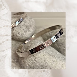 Puede incluir: Un brazalete de plata con marcas de contraste, apoyado sobre una piedra gris. El brazalete es redondo y tiene una superficie lisa y pulida. Las marcas incluyen el número "925".