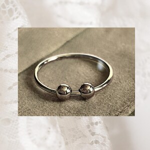 Puede incluir: Un anillo de metal plateado que presenta una banda circular delgada con dos pequeñas cuentas esféricas y lisas colocadas muy juntas en la parte frontal. El metal tiene un acabado pulido y reflectante que capta la luz de manera uniforme.