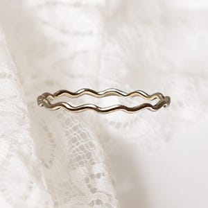 May include: A delicate gold ring with a wavy, textured design. The ring's unique shape adds a touch of elegance. The ring is set against a white, lacy background, highlighting its intricate details. This ring is a piece of jewellery.
