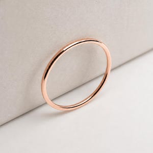 May include: A thin, circular ring crafted from rose gold-toned metal. The band features a smooth, polished surface with a consistent, rounded profile throughout its entire circumference, showcasing a simple and clean design without any added embellishments.