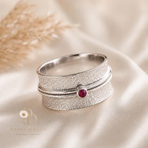 May include: A silver spinner ring featuring a wide band with a textured, hammered finish. The ring includes a thinner, smooth central band accented by a single, small, round, deep pinkish-red gemstone set in a polished silver bezel setting.