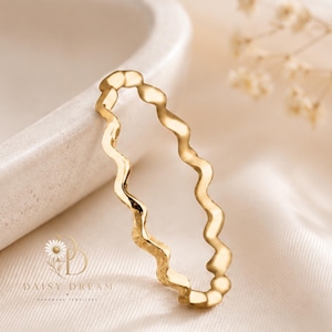 Puede incluir: Un anillo de tono dorado que presenta un diseño de banda continua y ondulada. La superficie metálica tiene un acabado liso y pulido que refleja la luz a lo largo de las curvas del anillo. El logotipo de Daisy Dream Handmade Jewellery es visible.