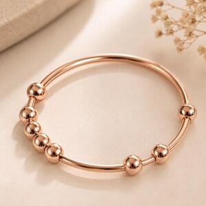 Puede incluir: Una pulsera rígida circular en tono oro rosa que presenta una banda de metal fina y lisa adornada con siete pequeñas cuentas esféricas pulidas, distribuidas a lo largo de la circunferencia del marco de alambre circular con un diseño simple.