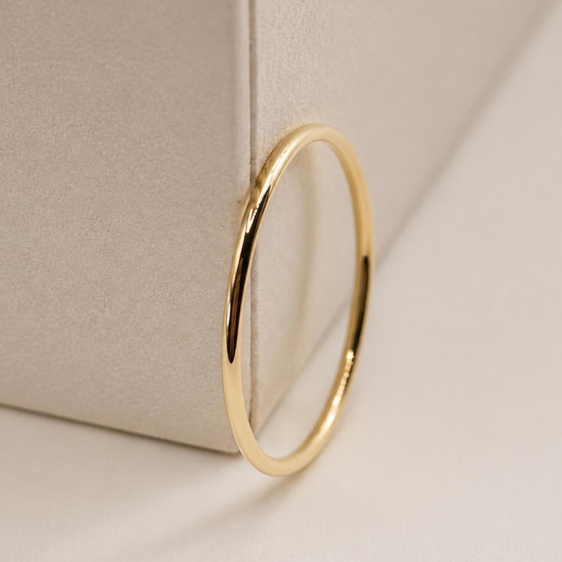 Puede incluir: Un anillo fino de oro amarillo pulido con una banda lisa y redondeada. El anillo presenta un dise&ntilde;o circular sencillo con una superficie met&aacute;lica reflectante que capta la luz, mostrando un aspecto limpio y uniforme en toda la banda.