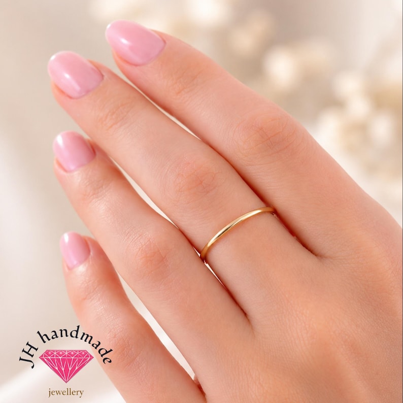 Puede incluir: Un anillo de banda de oro fino, liso y pulido, usado en el dedo anular. El anillo presenta un dise&ntilde;o circular simple con un acabado met&aacute;lico reflectante. El logotipo JH handmade jewellery con un icono de diamante rosa es visible.