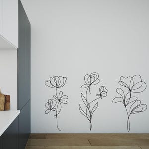 May include: Black line art floral wall decals. Three different floral designs are displayed on a white wall. The flowers have stems and leaves. The wall art is placed above a wooden floor.