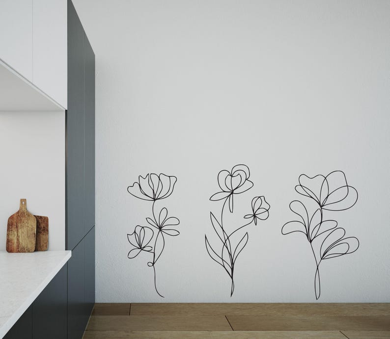 May include: Black line art floral wall decals. Three different floral designs are displayed on a white wall. The flowers have stems and leaves. The wall art is placed above a wooden floor.