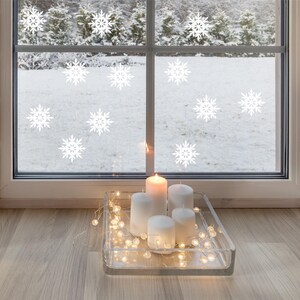 May include: A cozy winter scene with a window decorated with white snowflake decals. A clear glass tray holds white pillar candles and a string of fairy lights. The window overlooks a snowy landscape, creating a warm, inviting atmosphere.