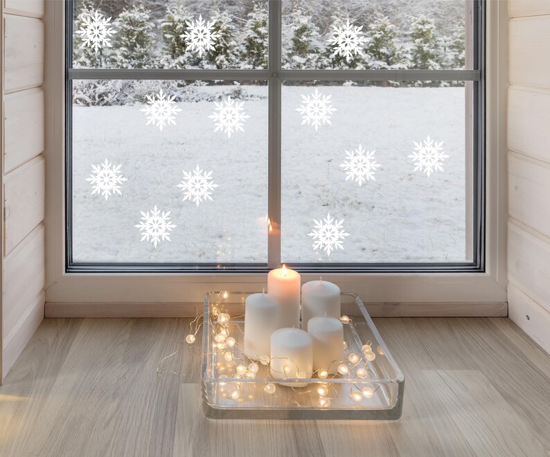 May include: A cozy winter scene with a window decorated with white snowflake decals. A clear glass tray holds white pillar candles and a string of fairy lights. The window overlooks a snowy landscape, creating a warm, inviting atmosphere.