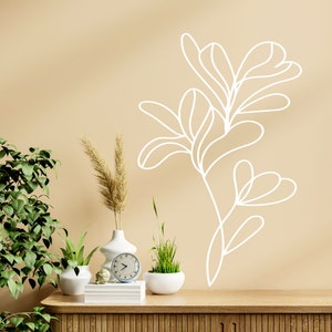 Op de afbeelding: Een beige muur met een witte lijntekening van een plant. Een houten plank met drie potplanten, een witte vaas met gedroogd pampasgras en een witte klok.