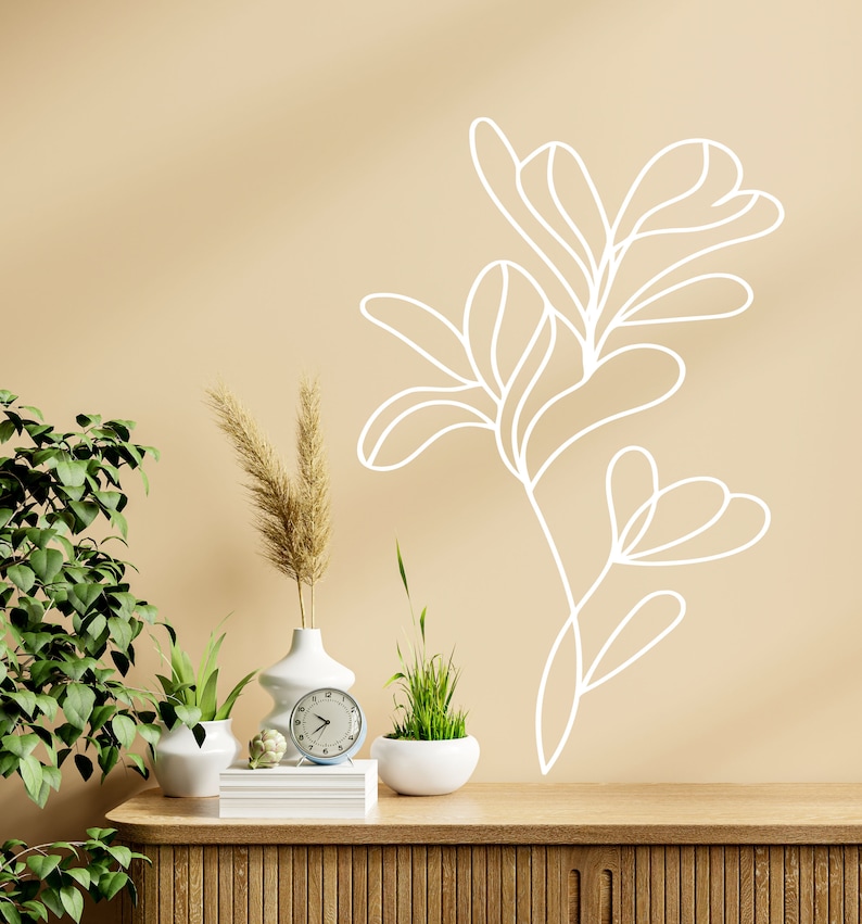 Op de afbeelding: Een beige muur met een witte lijntekening van een plant. Een houten plank met drie potplanten, een witte vaas met gedroogd pampasgras en een witte klok.