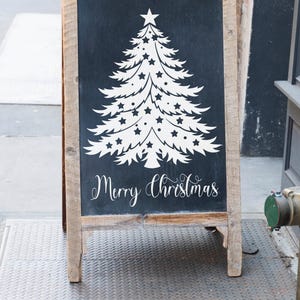 May include: A wooden A-frame chalkboard sign with a white silhouette of a Christmas tree decorated with stars and the text "Merry Christmas".