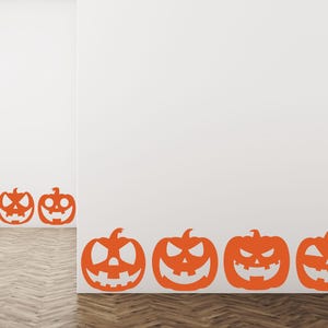 Puede incluir: Adhesivos de pared de calabazas de Halloween naranjas con caras talladas se muestran en una pared blanca. Las calabazas est&aacute;n dispuestas en fila a lo largo de la base de la pared, creando un ambiente festivo y espeluznante.