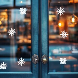 May include: A teal-colored double door with white snowflake decals. The door has ornate hardware and a blurred background of a warmly lit interior. The snowflakes are evenly spaced across the glass panels, creating a festive winter scene.