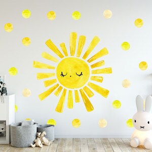 May include: A yellow watercolor sun with a smiling face and closed eyes, surrounded by yellow watercolor dots on a white wall. The sun is a wall decal and is part of a nursery decor set.