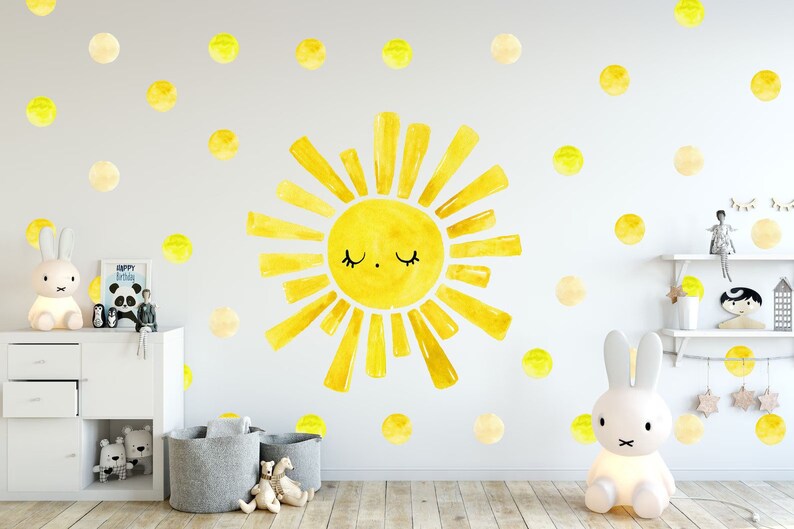 May include: A yellow watercolor sun with a smiling face and closed eyes, surrounded by yellow watercolor dots on a white wall. The sun is a wall decal and is part of a nursery decor set.