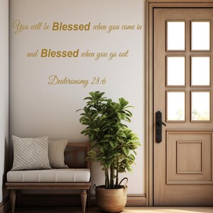 Pu&ograve; includere: Un adesivo da parete con il testo "You will be Blessed when you come in and Blessed when you go out Deuteronomy 28:6" in lettere dorate. L'adesivo &egrave; su una parete bianca con una porta in legno e una piccola panca con un cuscino.