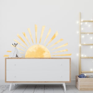 May include: A yellow watercolor sunburst wall decal with a white background. The decal is above a white dresser with two drawers.
