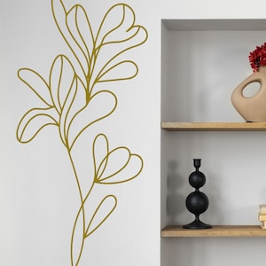 K&ouml;nnte beinhalten: Eine goldene Linienzeichnung einer stilisierten Pflanze mit Bl&auml;ttern an einer wei&szlig;en Wand. Die Pflanze befindet sich auf der linken Seite des Bildes und ein wei&szlig;es Regal mit einem schwarzen Kerzenhalter und einer braunen Vase mit roten Blumen befindet sich auf der rechten Seite des Bildes.
