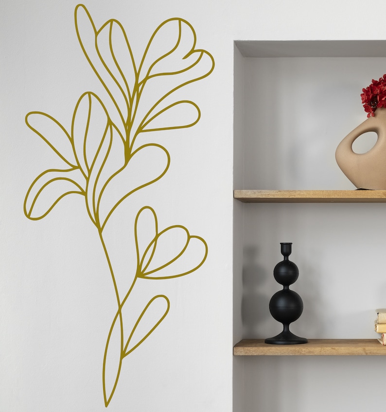 K&ouml;nnte beinhalten: Eine goldene Linienzeichnung einer stilisierten Pflanze mit Bl&auml;ttern an einer wei&szlig;en Wand. Die Pflanze befindet sich auf der linken Seite des Bildes und ein wei&szlig;es Regal mit einem schwarzen Kerzenhalter und einer braunen Vase mit roten Blumen befindet sich auf der rechten Seite des Bildes.