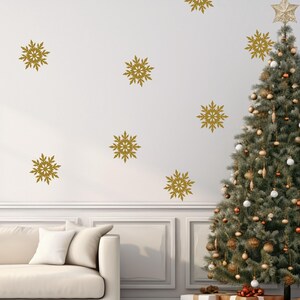 May include: A decorated Christmas tree with gold ornaments and a star topper stands next to a white wall adorned with gold snowflake decals. A white couch and wrapped gifts complete the festive scene.