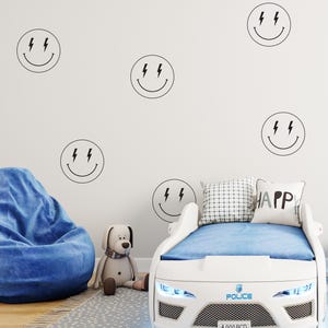 May include: White car-shaped bed frame with blue bedding and a blue and white pillow with the word "HAPPY" on it. The bed frame has a police emblem and the text "POLICE" and "A 000 BCD" on the front. The wall behind the bed has several black and white smiley face stickers with lightning bolts for eyes.