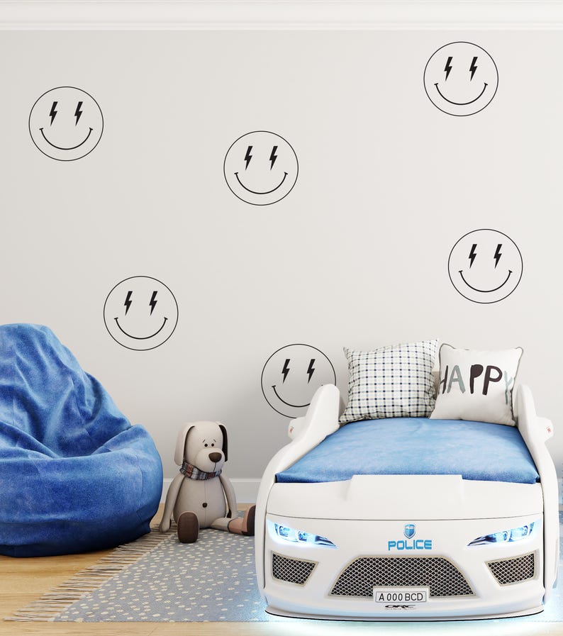 May include: White car-shaped bed frame with blue bedding and a blue and white pillow with the word "HAPPY" on it. The bed frame has a police emblem and the text "POLICE" and "A 000 BCD" on the front. The wall behind the bed has several black and white smiley face stickers with lightning bolts for eyes.