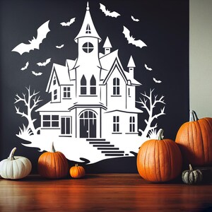 May include: A white haunted house silhouette with bats and bare trees is displayed on a black wall. Several orange pumpkins and one white pumpkin are arranged on a wooden surface, along with a decorative metal container.