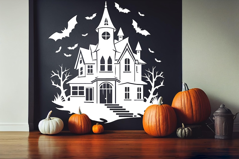 May include: A white haunted house silhouette with bats and bare trees is displayed on a black wall. Several orange pumpkins and one white pumpkin are arranged on a wooden surface, along with a decorative metal container.
