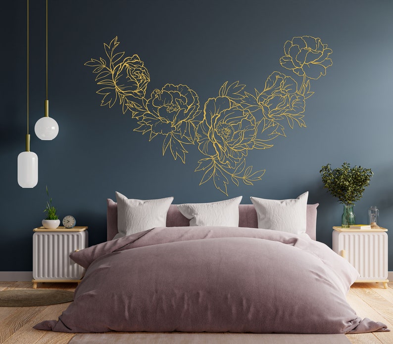 May include: A bedroom with a dark blue wall featuring a gold floral design. The bed has a light pink duvet and white pillows. There are two white nightstands with lamps on either side of the bed.