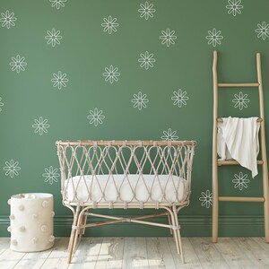 May include: A white wicker crib with a white mattress, a white pom-pom storage bin, and a wooden ladder leaning against a green wall with white daisy decals.
