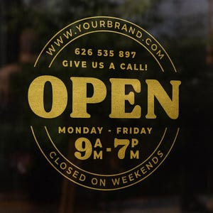 May include: Gold-colored "OPEN" sign on a dark background. The sign includes the website address "WWW.YOURBRAND.COM", a phone number, and the hours of operation: Monday to Friday, 9 AM to 7 PM. The sign also states "CLOSED ON WEEKENDS".