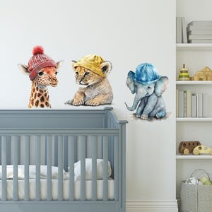 May include: Three watercolour animal wall decals with hats. A giraffe wearing a red beanie, a lion wearing a yellow beanie, and an elephant wearing a blue beanie.