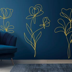 May include: Gold line art floral wall decals on a dark blue wall. The decals depict stylized flowers and leaves. A blue velvet armchair and a potted plant are in the foreground, creating a modern interior design.