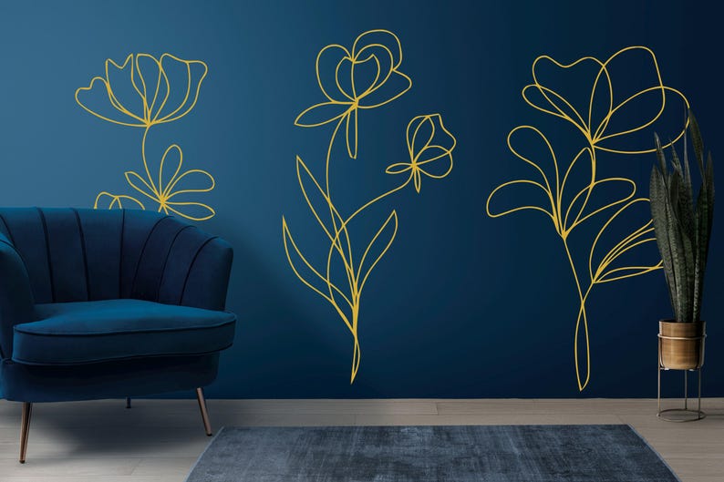 May include: Gold line art floral wall decals on a dark blue wall. The decals depict stylized flowers and leaves. A blue velvet armchair and a potted plant are in the foreground, creating a modern interior design.